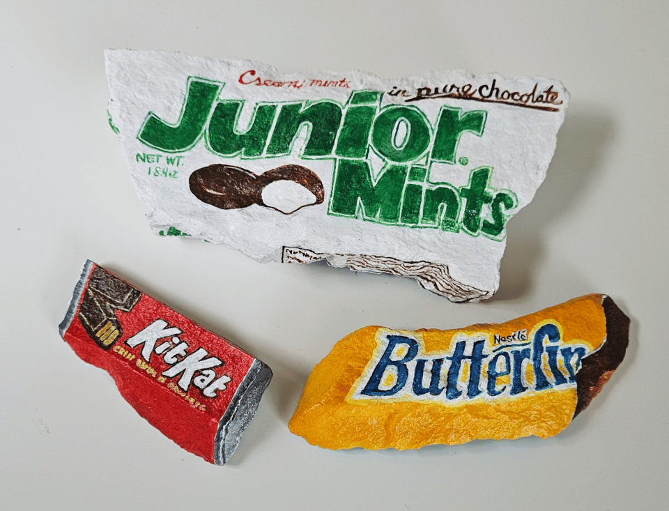 Three rocks painted to look like a box of Junior Mints, a Kit Kat, and a Butterfinger bar. They looks slightly crumpled and deformed, as if they were the original real packages and they'd been left out in the rain