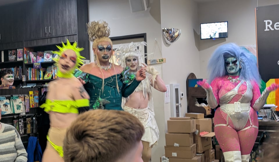 Five drag performers with colorful makeup and wild costumes dance in a bookstore in front of a rapt audience