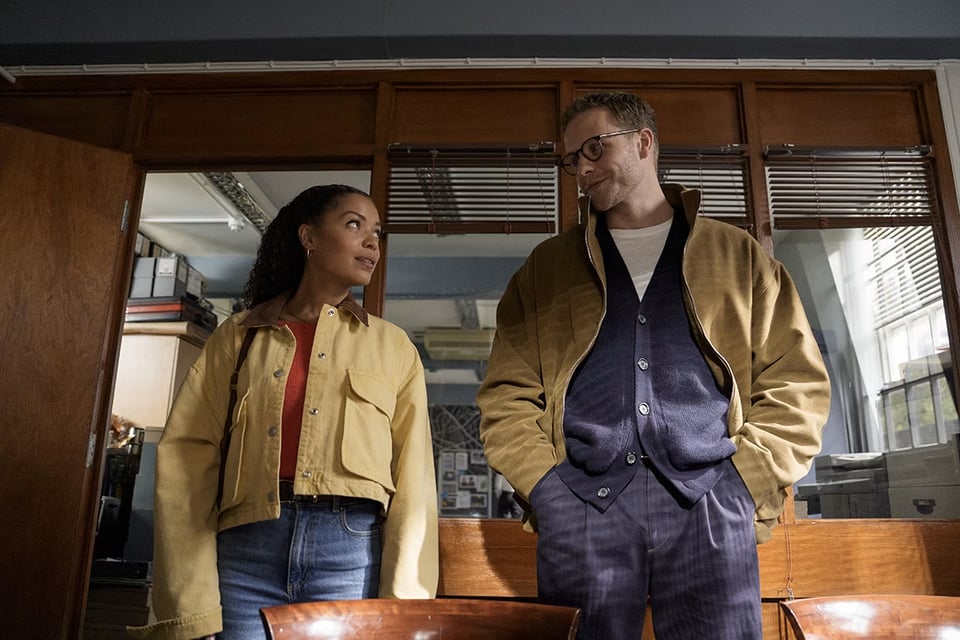 A man and a woman stand in an office and give each other a knowing look.