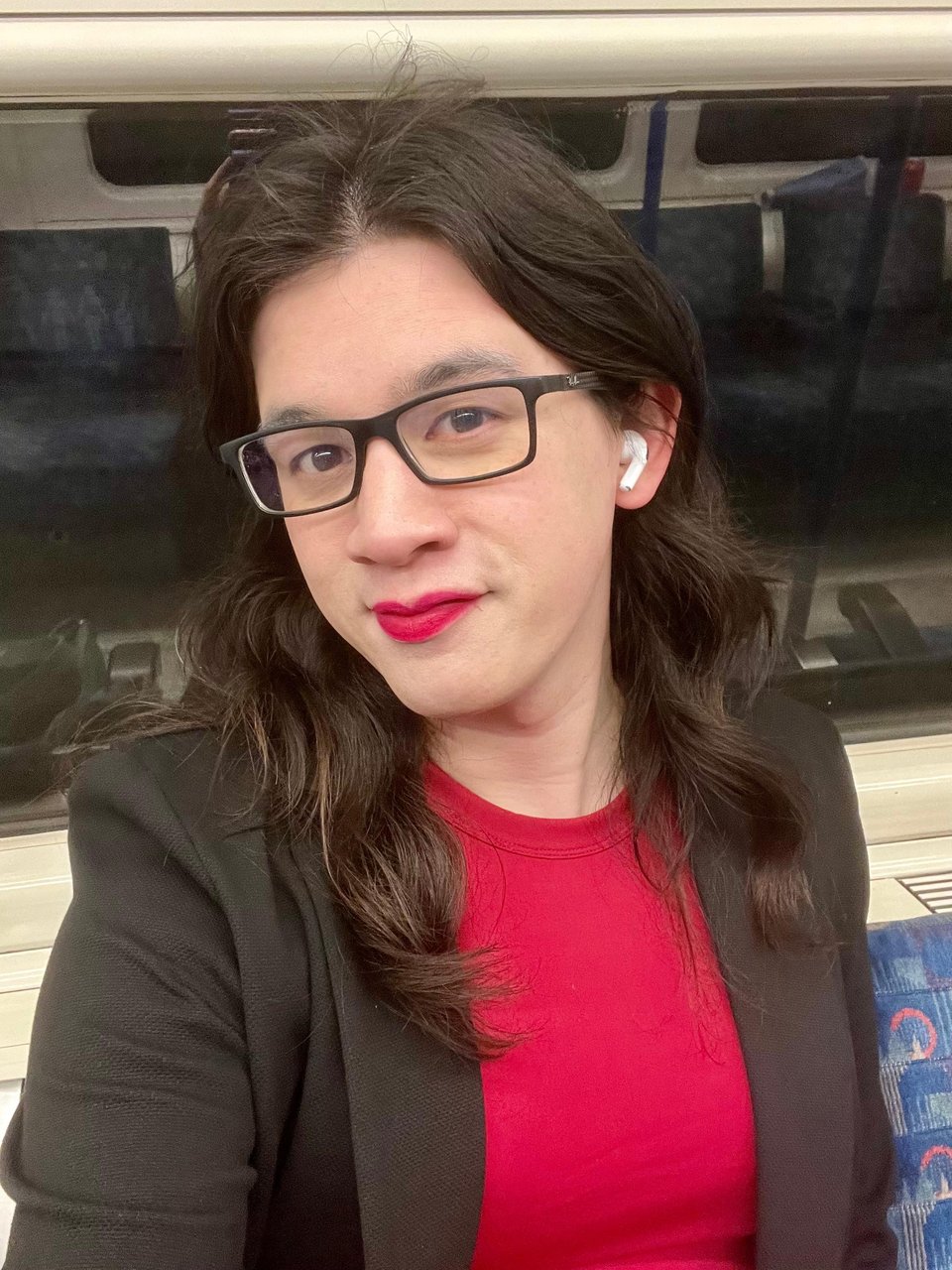 A selfie! I'm smiling at the camera while sitting on a Tube train. I have light-coloured skin, black rectangular glasses, and long brown hair falling on waves on either side of my face. I'm wearing bright red lipstick that matches my dress, and a black blazer.