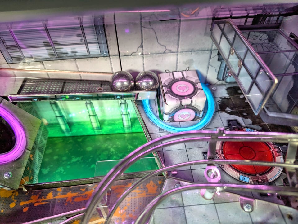 A close look inside parts of the P3 Portal pinball machine. You see two balls lying next to a Companion Cube. On the very left side is a pink portal.