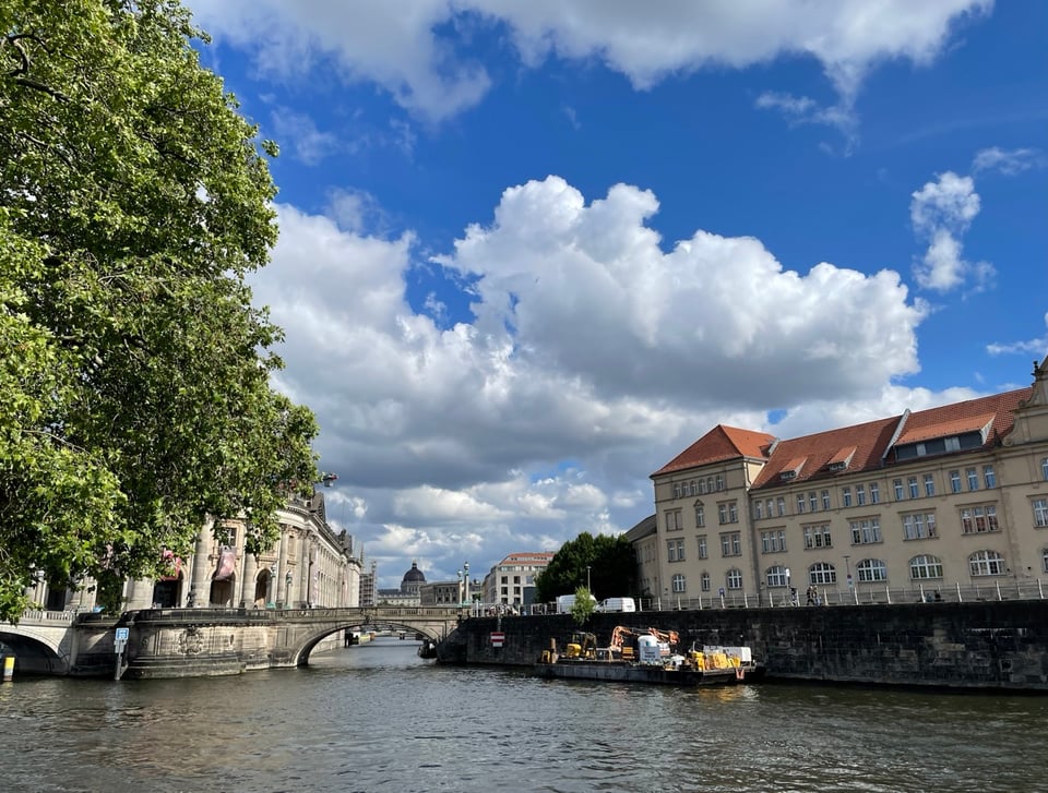 A view of the spree river looking from the side of Ebertbrücke towards Bode Museum. There is a tree in the foreground. Blue skies with clouds in the middle in the background and buildings on the edge of the photo
