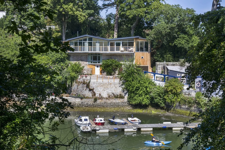 This alt text will take a minute because this is a complicated building to describe. It's sitting on a quayside in a creek and set into a cliffside. The lower two storeys are an old stone kiln building with a modern door set in the ground floor and a 1960s window set into the middle floor. To one side, a new wooden tower has been built with a garage on the ground floor and windows at the top, accessing the third floor of the original building. This third floor is a replica of the 1960s building, using wood and glass to create a rather Californian look with a zigzag roof and glass curtain walls.