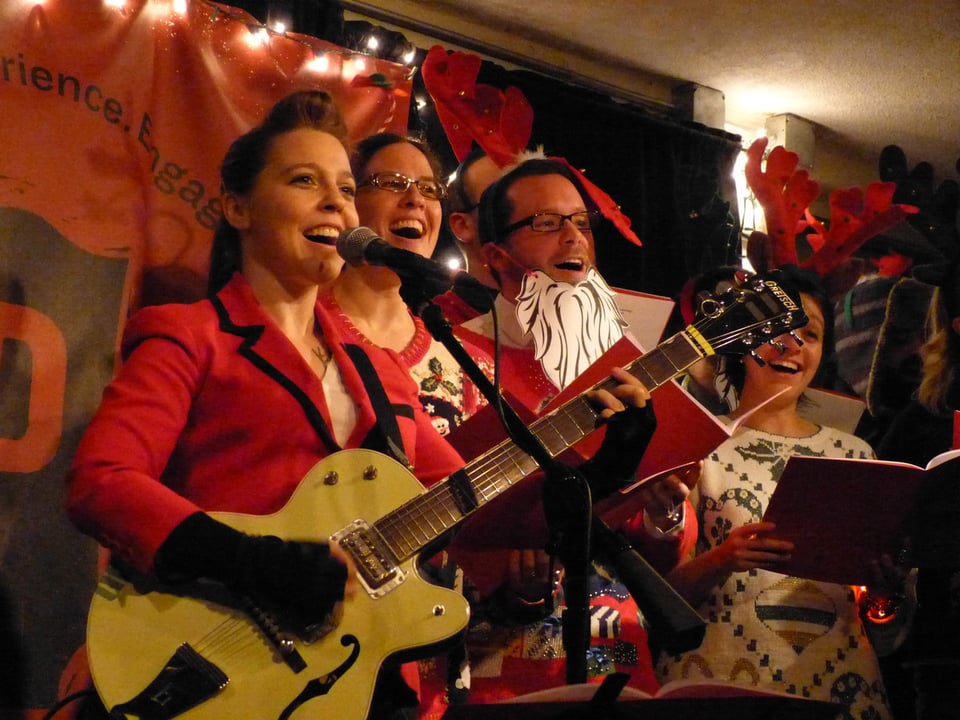 A woman with a yellow semi-hollowbody electric guitar stands at a microphone, with three singers wearing Christmas-y outfits standing to her left.