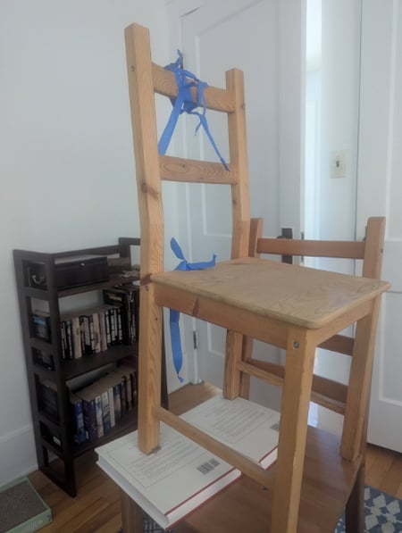 A stack of two wooden chairs, the upper one tilted using a pair of books and festooned with painter's tape