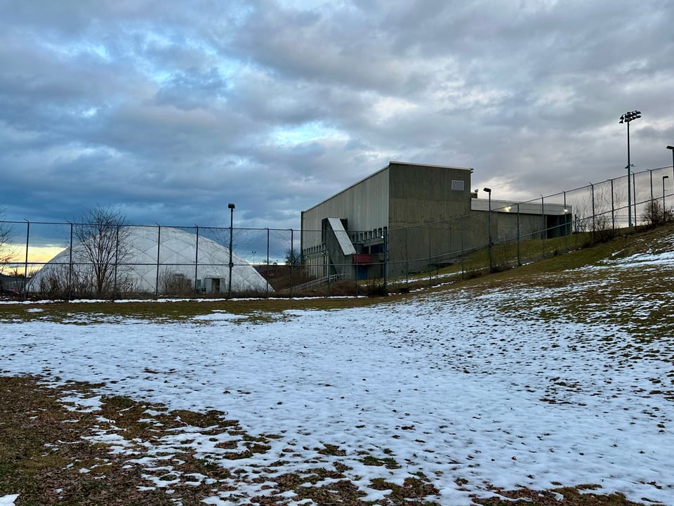 photo of windowless concrete building and white fabric dome, behind a 20' high chain-link fence, with snow on the grass in the foreground and a cloudy gray sky above