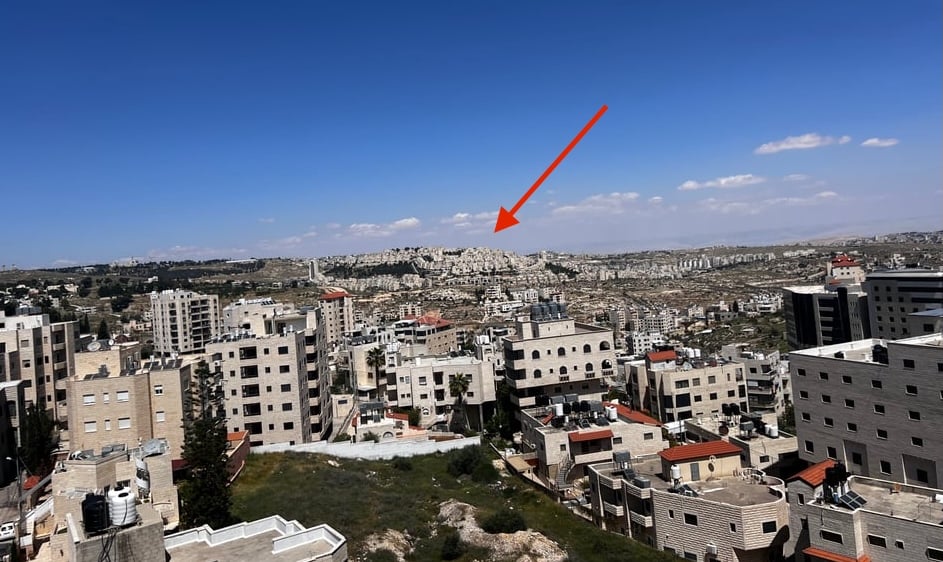 A view out a window of concete buildings with windows and a blue sky with a few clouds. A red arrow points to a collection of buildings, appearing to be a town, in the distance