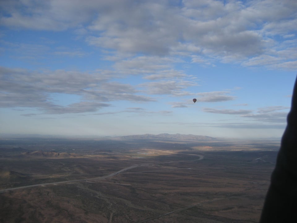 Open sky, clearly at altitude, above a crinkled brown desert. The small dot of a hot air balloon is just visible.