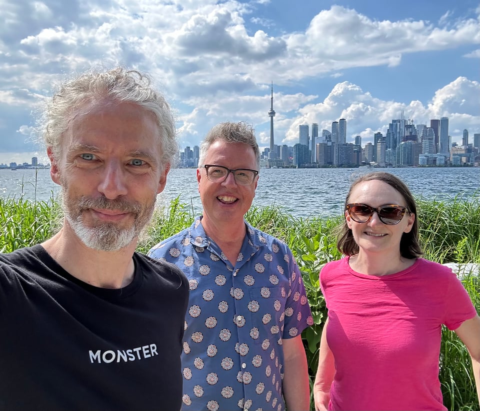 Photo of 3 white people standing by the high grass on Algonquin Island, with the mainland of Toronto in the background