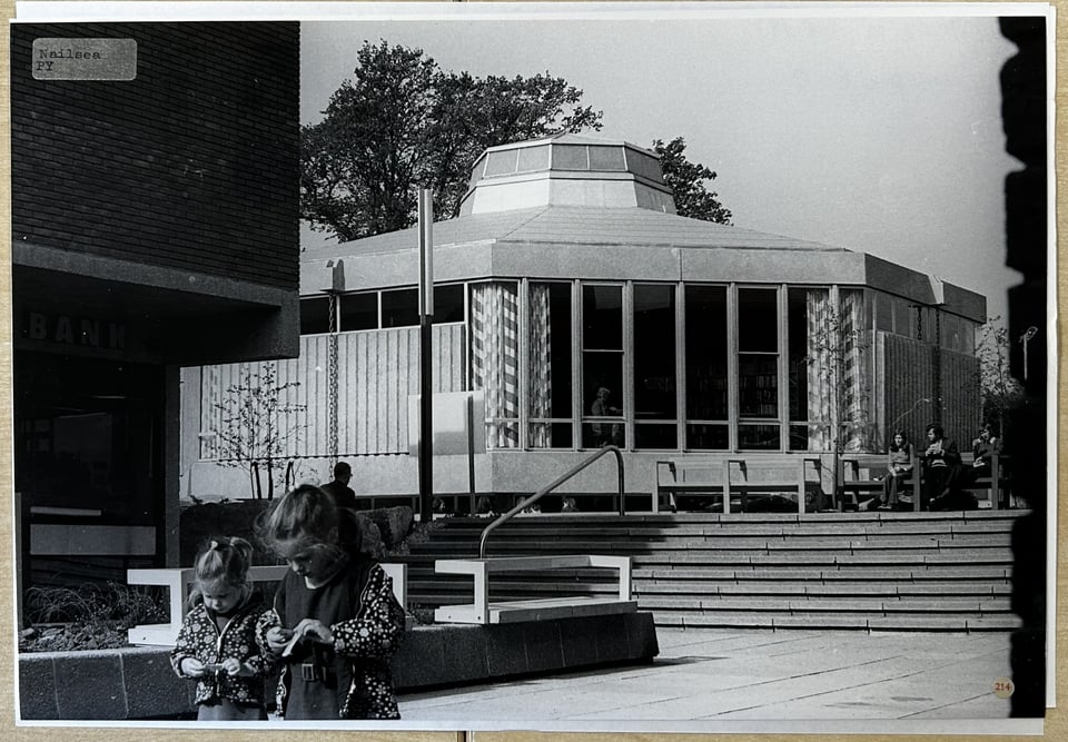 A black and white photo of the library exterior from the shopping centre. It's octagonal shape along with the lantern on it and the gap created by the jettied upper ground floor make it look like a spaceship. If spaceships had zigzag print curtains. In the foreground, two young girls in floral quilted coats and walking.