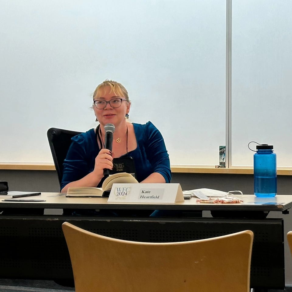 Kate has her blond hair pulled back and is wearing a dark teal velvet dress. She is holding a microphone, sitting behind a desk and readnig from a book while looking out at the audience..