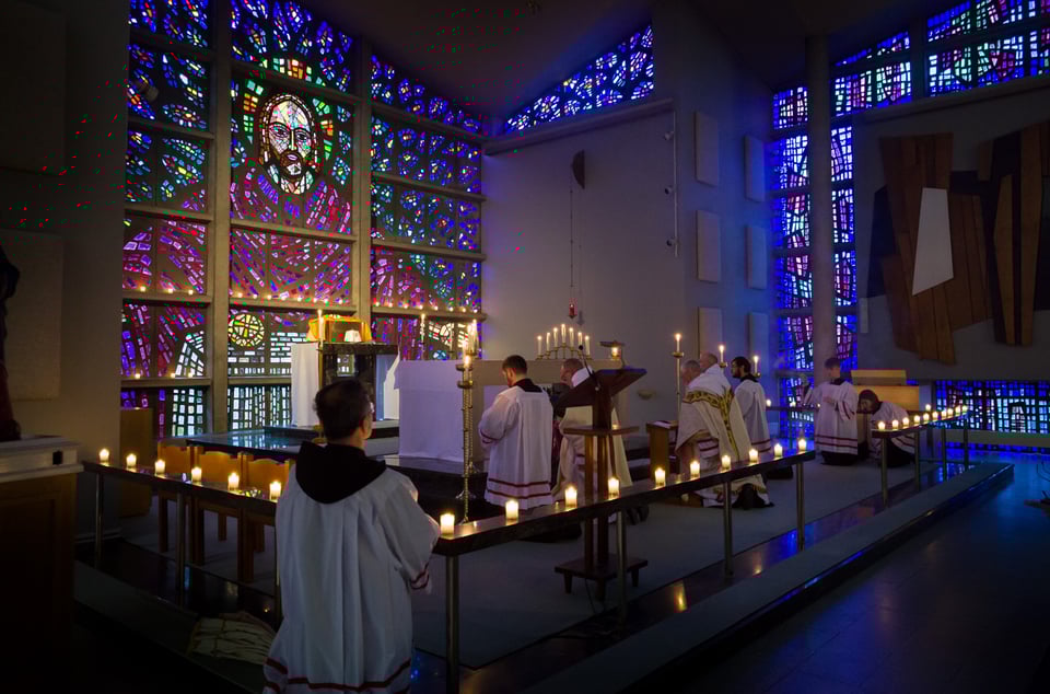 A wall of glass mosaic showing Jesus Christ blessing the bread and wine. It glows with blues, greens and purples, completely dominating the actual priests taking a candlelit service in front of it. The side wall of the chapel is also blue and green dalle de verre.