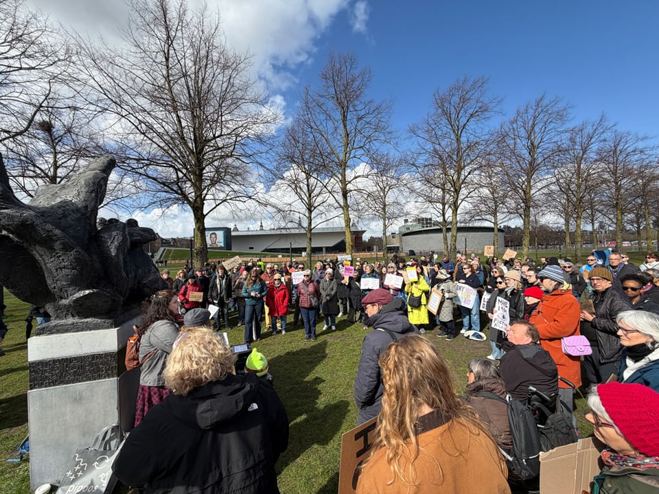 A crowd of people stand in a large circle around people speaking at a protest in Amsterdam.