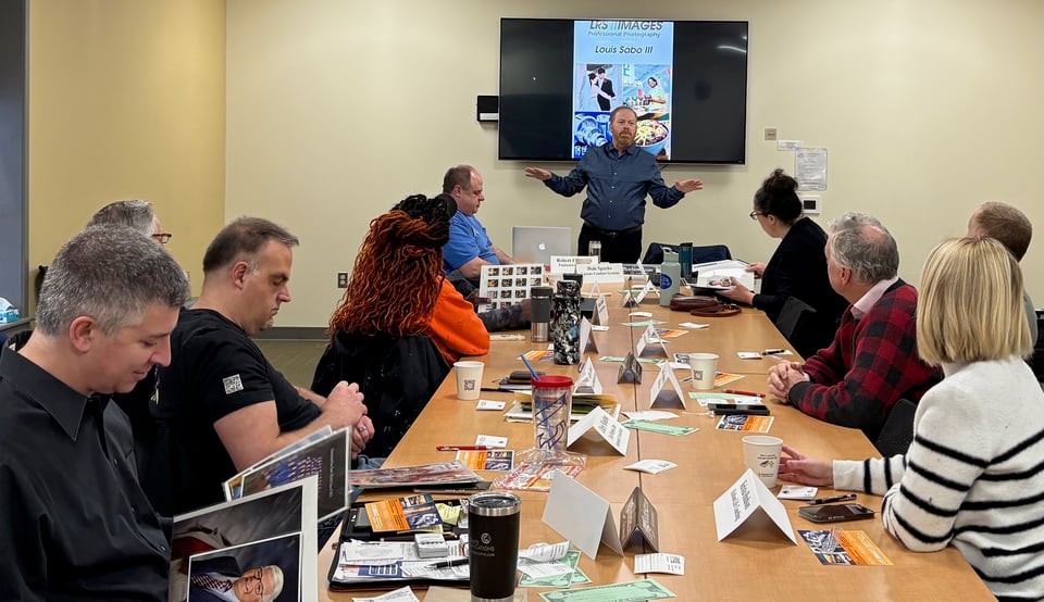 Lou Sabo stands in front of NRG members, presenting to them with a wall-mounted TV display showing his photos behind him.