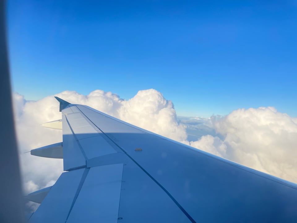 The left wing of an airplane flying over puffy white clouds with blue sky overhead.