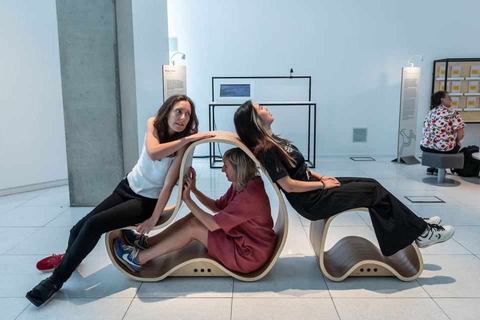 three people in a white gallery space sit on and inside a hollow wooden sculpture