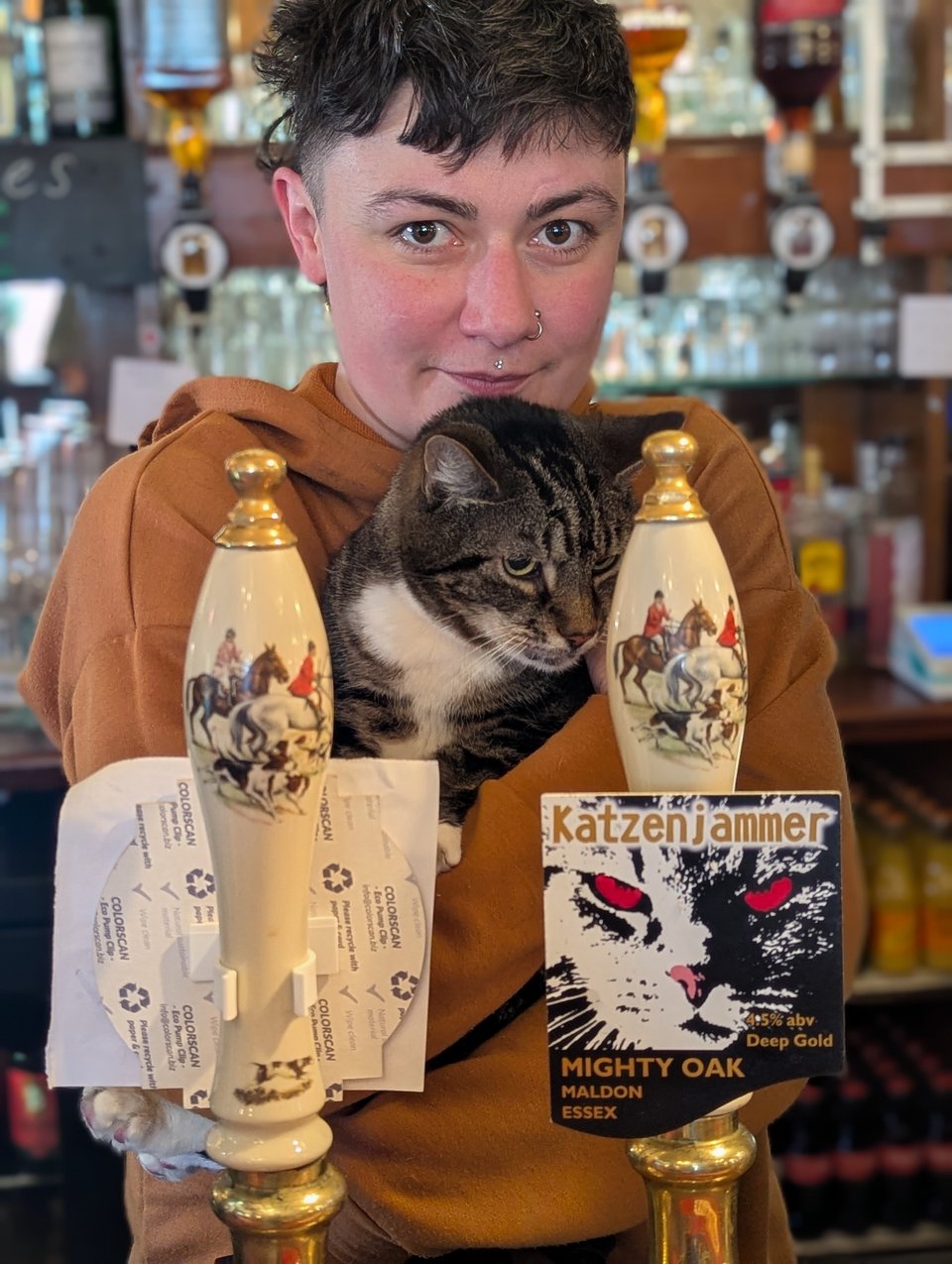 me looking frankly quite deranged because I forgot to ask the customer taking the photo to make me look nice, holding up my big tabby cat next to the Katzenjammer pump clip, which features a tabby cat with glowing red eyes