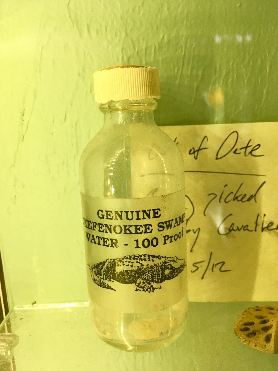 a small jar on a shelf. its label shows an alligator and reads "genuine Okefenokee Swamp Water - 100 Proof."