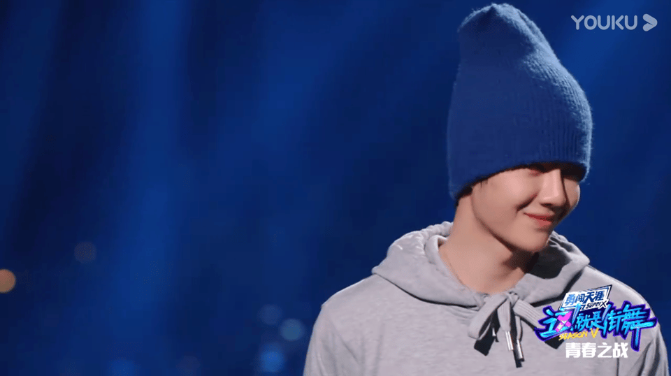 screencap from the finale of Street Dance of China season 5. Against a royal blue background, Wang Yibo stands towards the right of the photo in a grey sweater with hoodie, wearing a classic blue fleece b-boy toboggin the same shade of bright royal blue as the stage lighting. He's looking off to his left side and his eyes are almost completely shadowed by his cap, which comes down over the top half of his face. The bottom half of his face, however, is glowing. He is smiling a secretive, sweet small smile, and his cheeks are round.