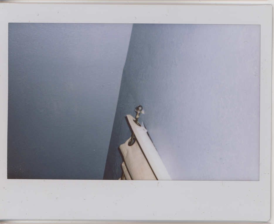 polaroid photo of a bare ceiling where it meets the wall and the corner of a metal curtain rod with cream curtains hanging from it