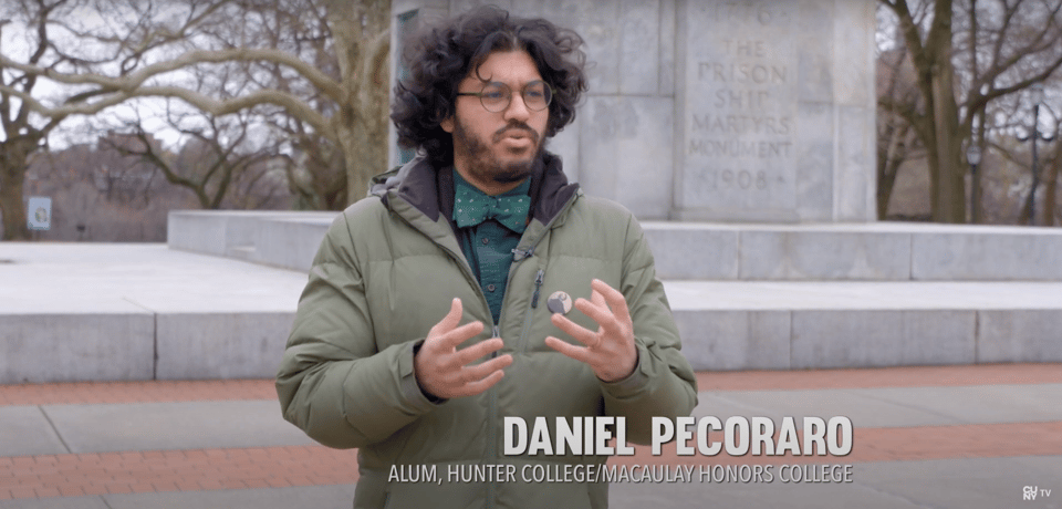 Daniel Pecoraro (that's me), a male-presenting person, with a green coat, dark green bowtie, and dark green shirt, gesturing in front of a monument.