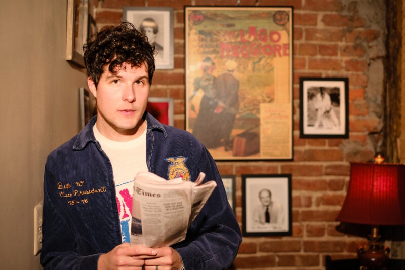 A man with dark, curly hair looks into the camera while holding a folding copy of the New York Times in his hands. Behind him is a brick wall with a poster surrounded by framed photographs.