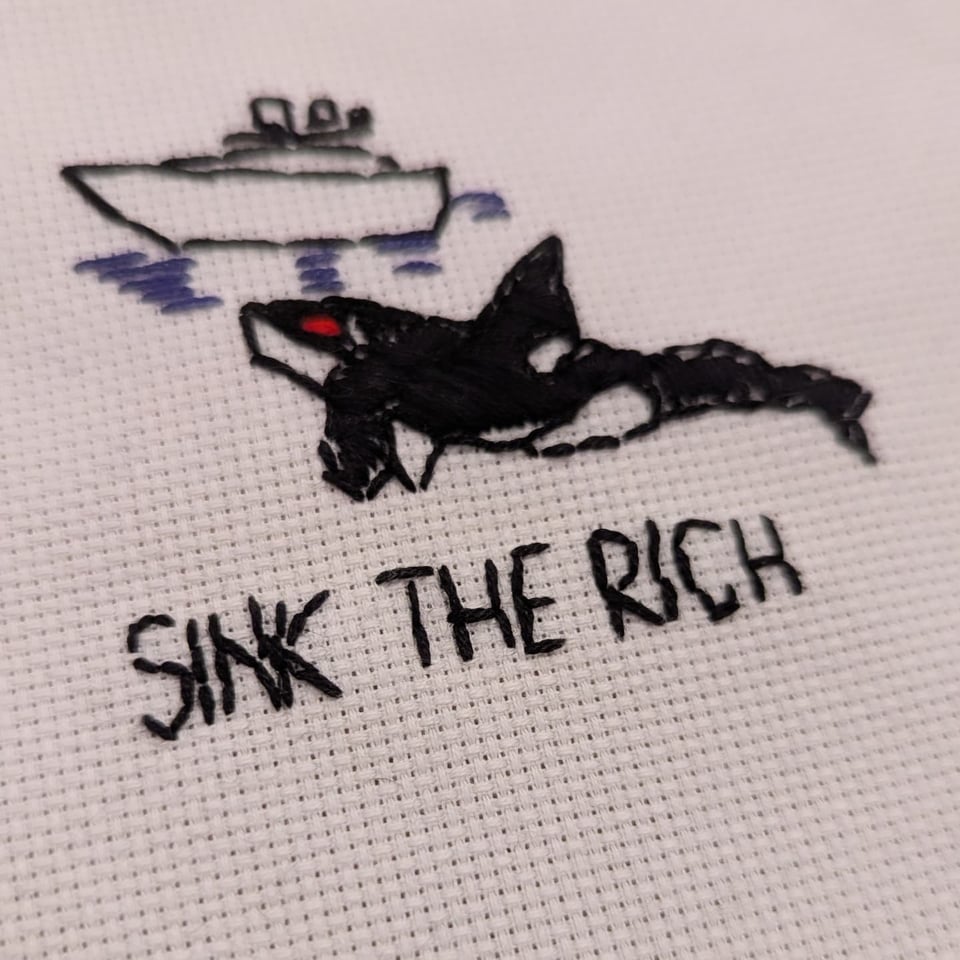 a piece of embroidery that features a yacht and a wobbly orca with a red eye coming to get it. The words sink the rich is below them.