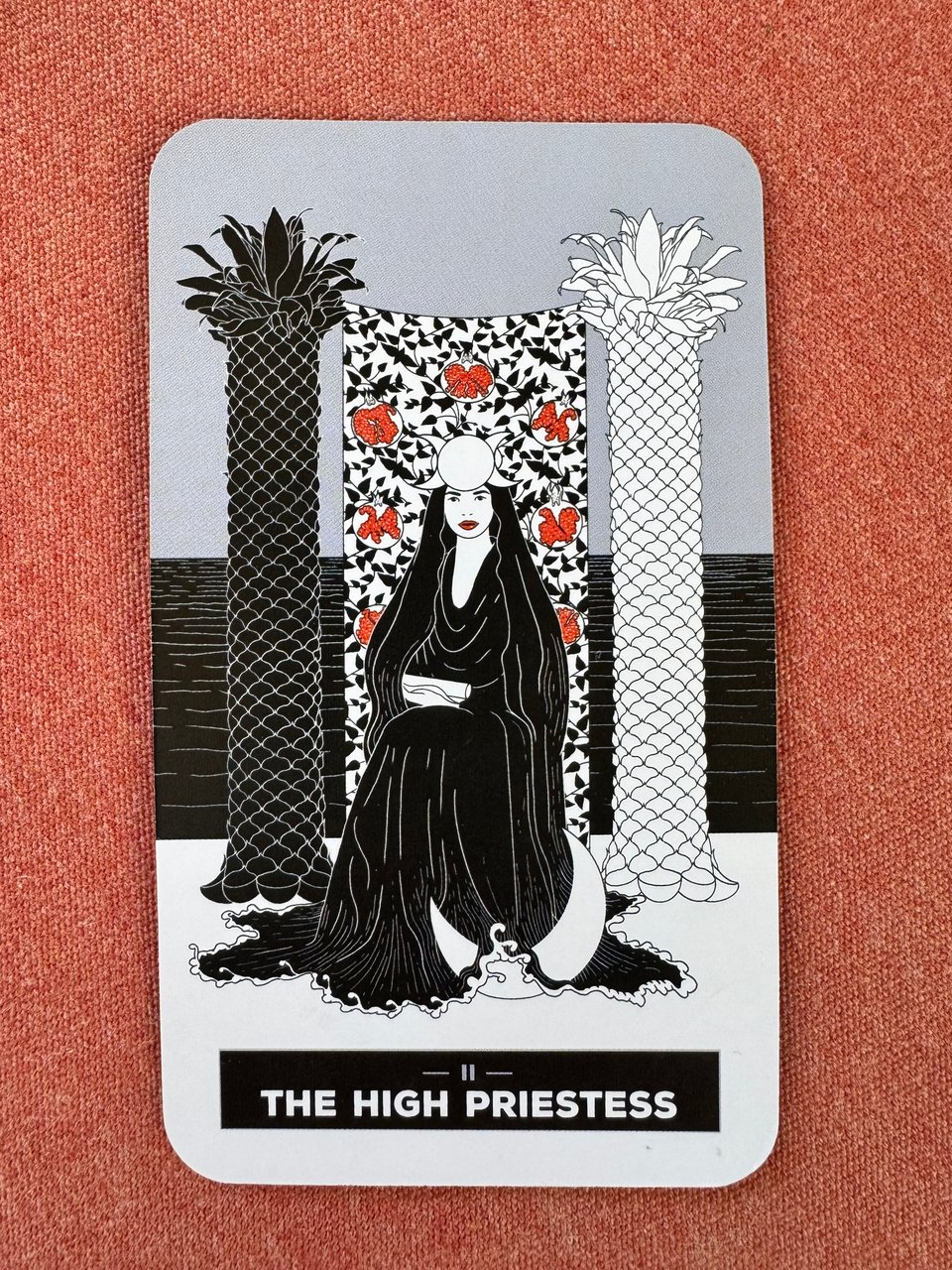 Tarot card depicting "The High Priestess" with a woman in a black robe and veil seated between two ornate pillars and a tapestry with pomegranates.
