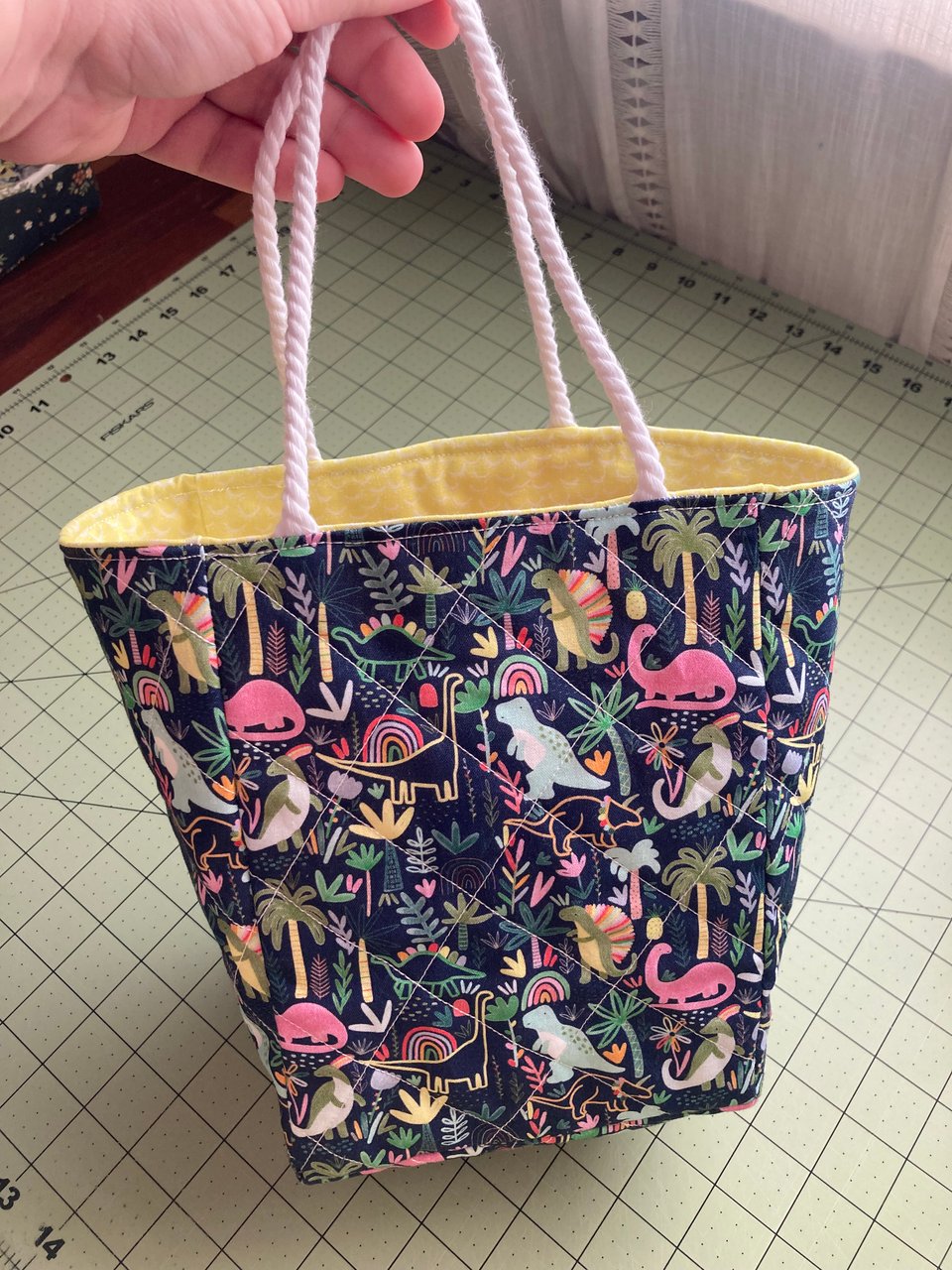 A small, quilted gift bag using a dark blue fabric decorated with a pattern of colorful dinosaurs and plants. The lining of the bag is a bright yellow with white scales.