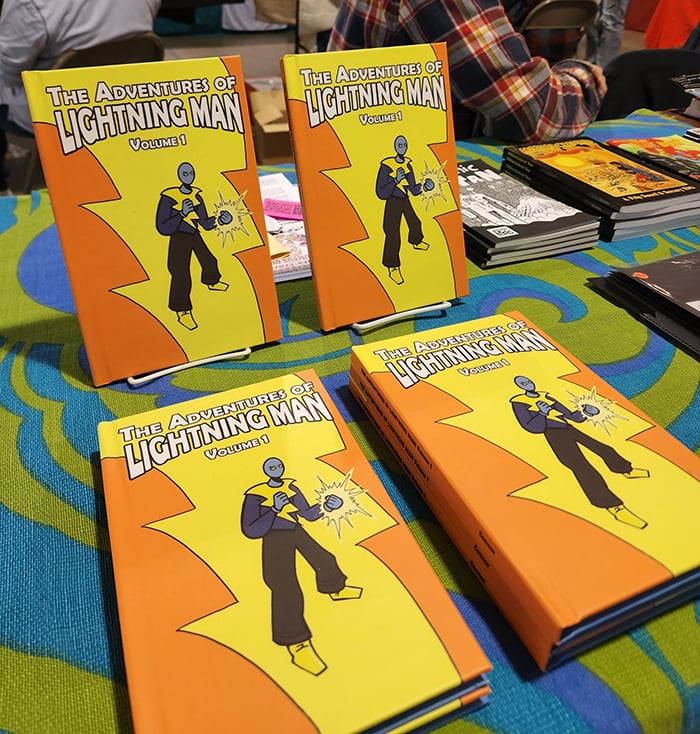 Several copies of the Lightning Man hardcover proudly displayed on my table at SPACE. Each cover features the heroic figure of Lightning Man, a guy in a costume, whose hand is crackling with electricity.