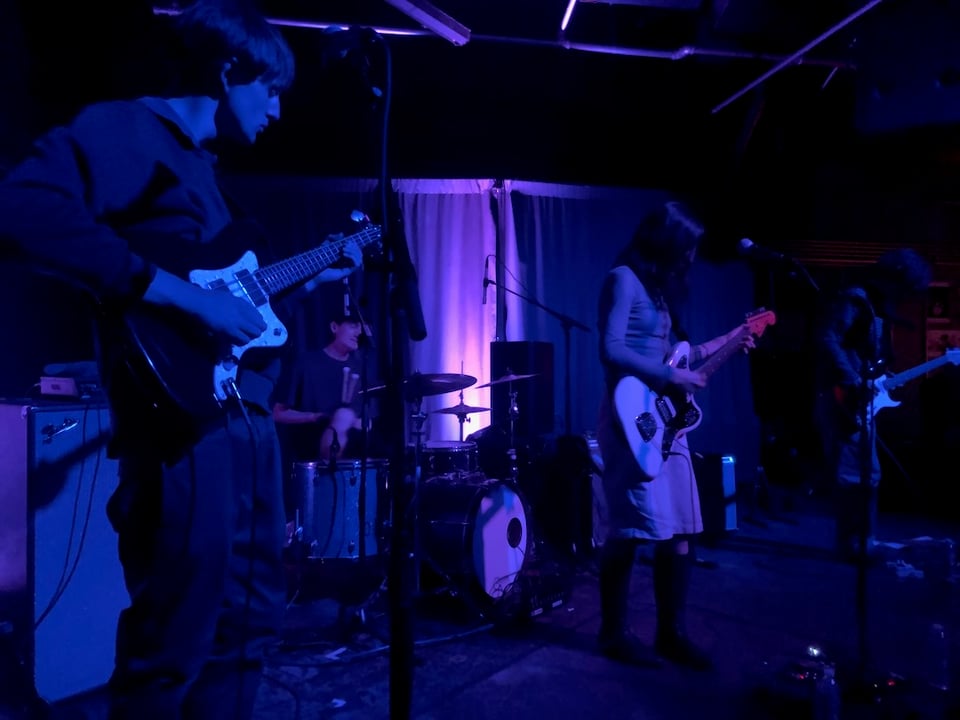Photo of Maria Somerville live at Zebulon. Stage is darkly lit. Maria Somerville stands in the center with head down, playing guitar there is another guitarist and a bassist, as well as a drummer in the photo.