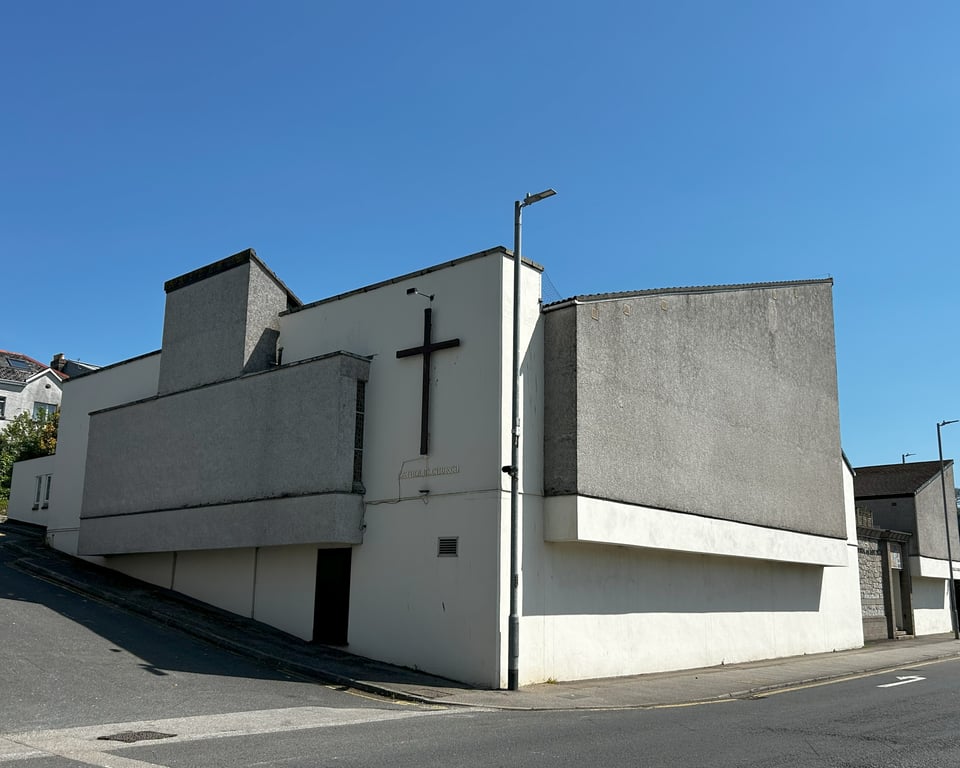 A large grey block of a building that appears to be materialising through the white walls of another large block. There's a simple cross on one white wall, and a very annoyingly placed lamppost.