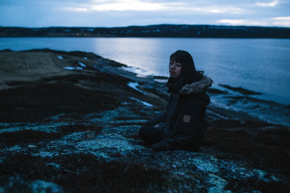 Under a dim sky, seated on a rock, facing away from the ocean.
