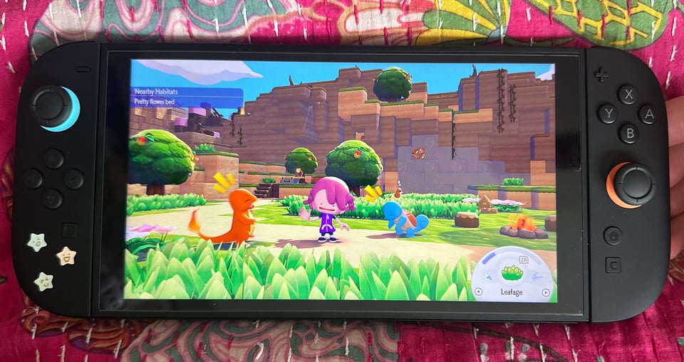 A switch 2 showing a still from Pokopia, where a Ditto pretending to be a purple haired girl is hanging out with a Squirtle and a Charmander