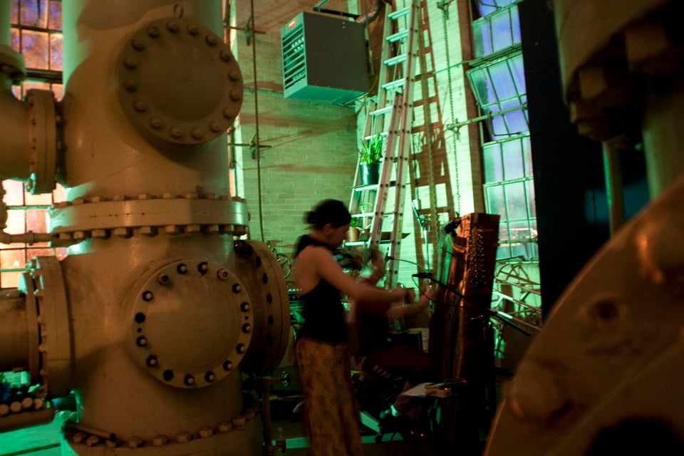 A woman playing violin and a man playing the inside of a piano vertically as a percussion instrument surrounded by pipes