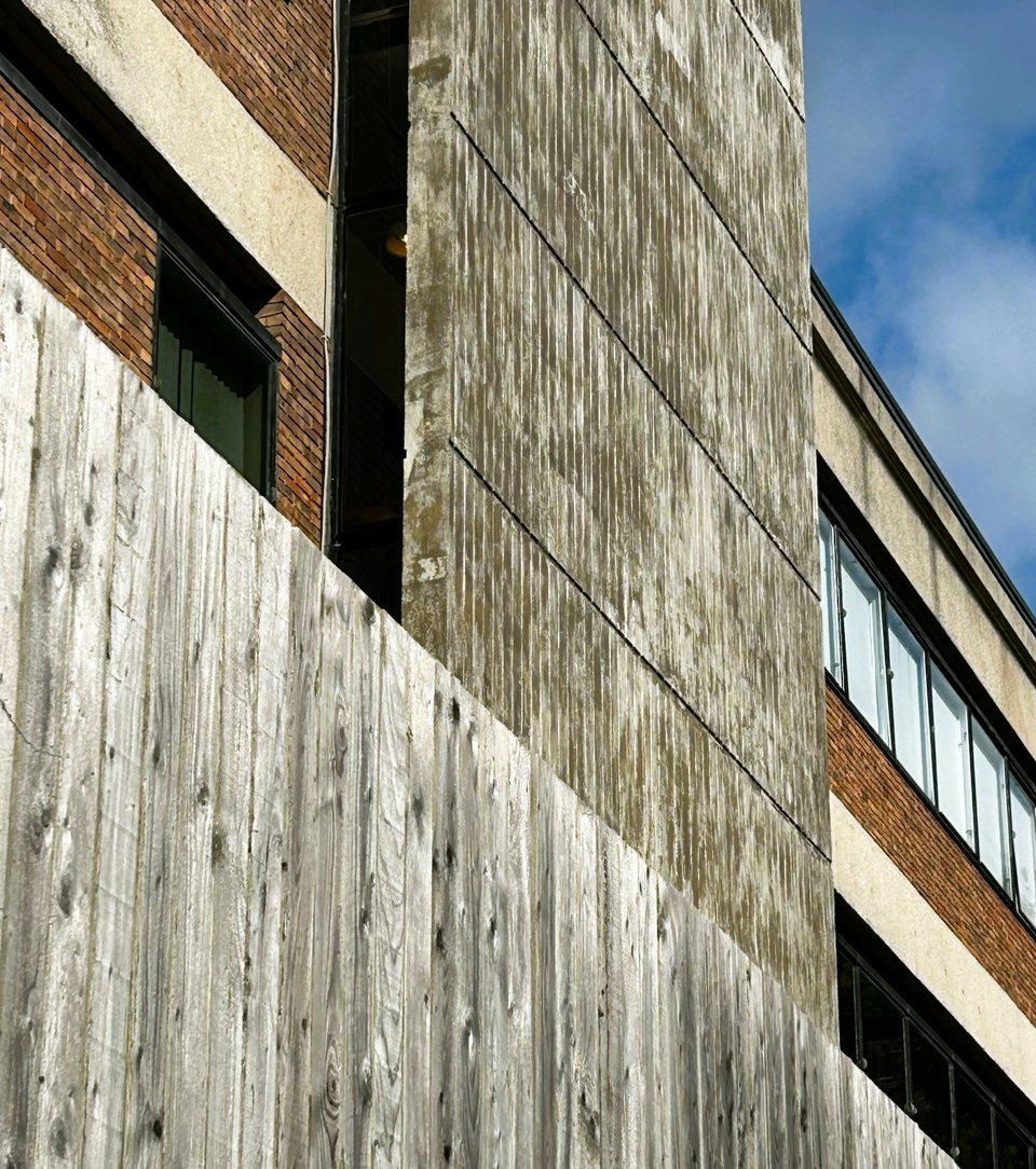 A wall of shuttered concrete rises several storeys high. The shutters had been made with the planks running vertically. In front of it is a wooden fence of vertical wood. Both elements are the same grey.
