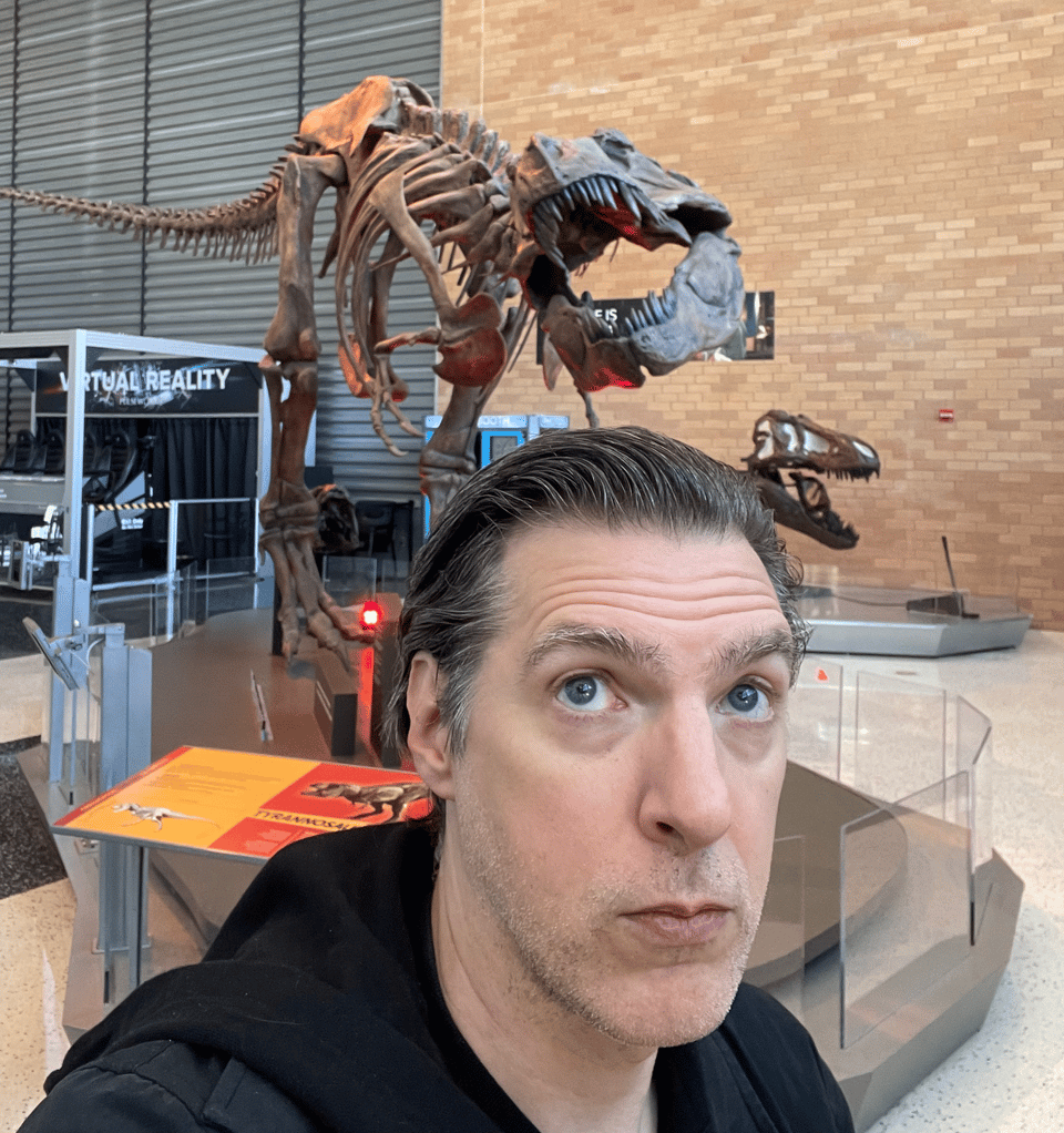 A tired man looks above him and sees the skeleton of a T-Rex in a museum lobby