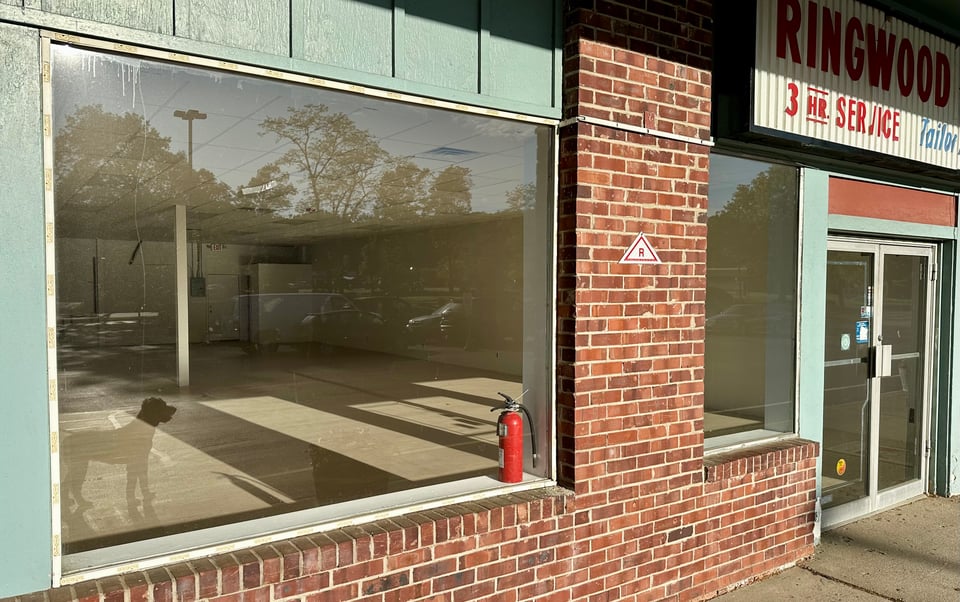 photo of abandoned dry cleaner, with brick and glass in front; the early morning sunlight is casting long shadows in the empty space; the parking lot and a dog are reflected in the glass