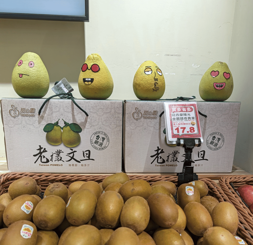In the foreground are a basket of kiwis like in a grocery store. In the background are two boxes of pomelos. Above the boxes are four pomelos that have been drawn on to look like cartoon characters.