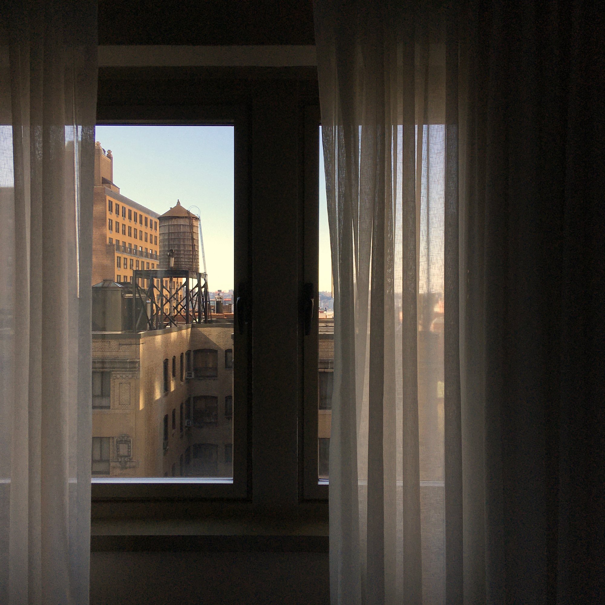 Photo out of a window view from a tall building, with curtains draped loosely on both sides of the window. The view across is of another building and its rooftop.