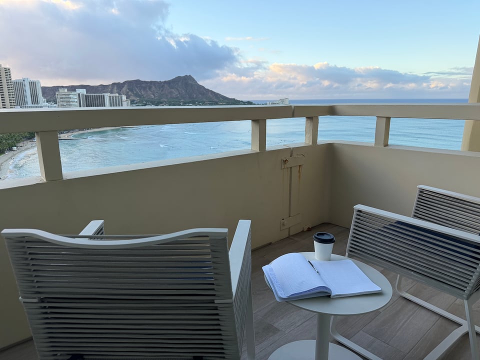 A view from the 21st floor of a hotel on Waikiki Beach.  A lanai with two chairs, a small round white table, and on the table, an open notebook and a paper cup of coffee with a plastic lid.