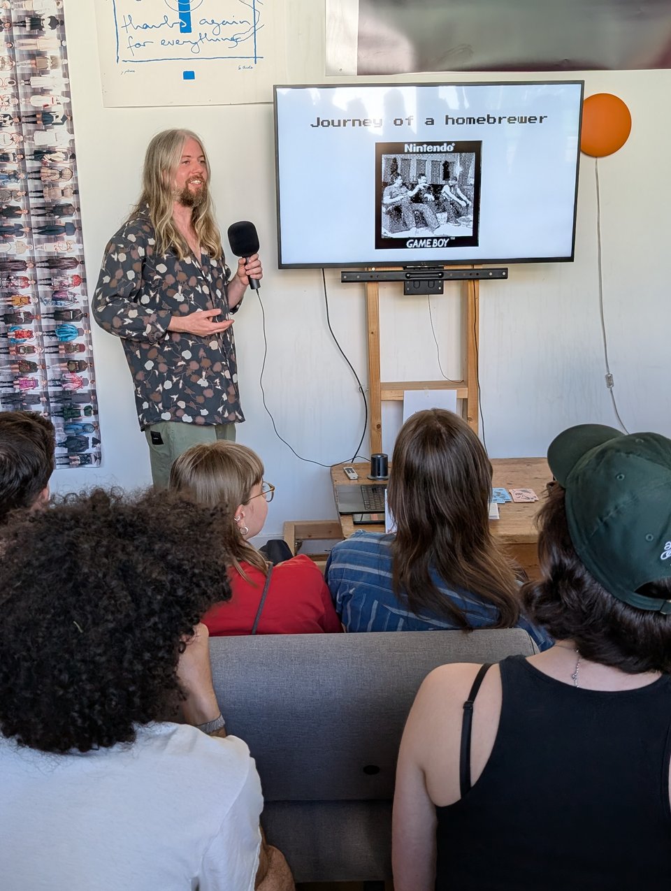 A long-haired man in a beautiful batik-adjacent shirt talks to an audience. The screen next to him says "journey of a homebrewer"