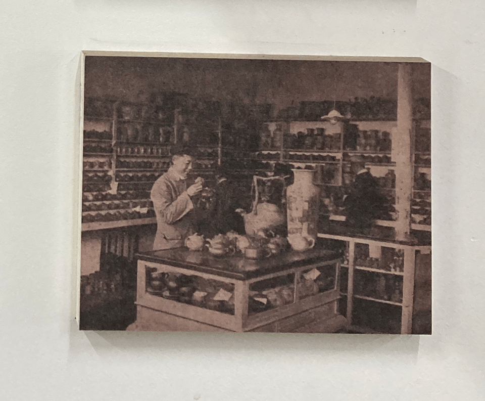 A man in a small shop surrounded by clay teapots. in the middle table sits a few large vases. he is inspecting a teapot by hand