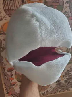 A puppet head, with light blue fleece skin and an open red mouth. Behind, a glimpse of the seasons of Wanderhome can be seen.