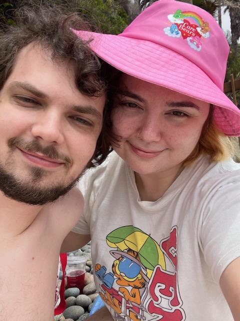 David Mariotte and his partner, Rebecca Ann, take a selfie on the beach. Rebecca has a summer Garfield shirt and a pink Care Bears bucket hat.