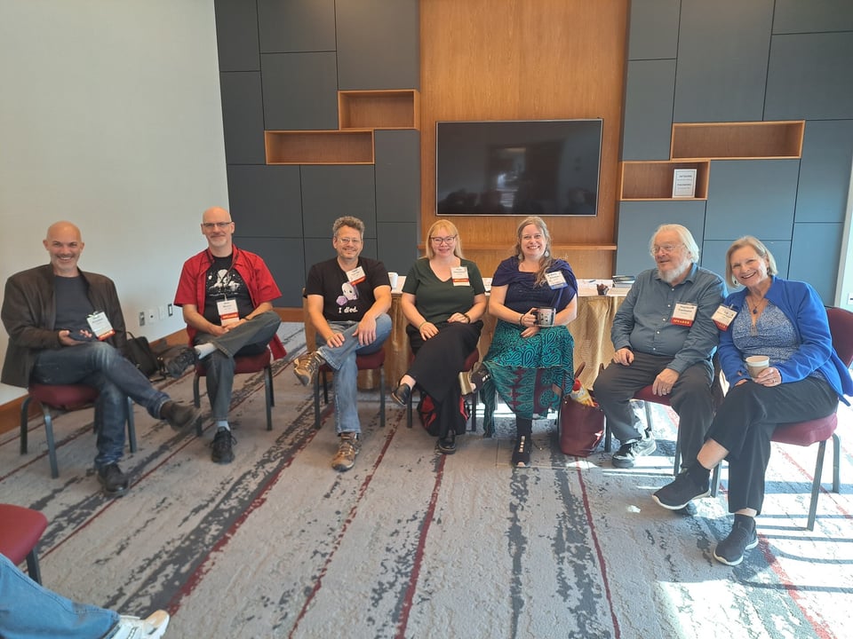 Seven smiling writers in chairs in a convention program room.