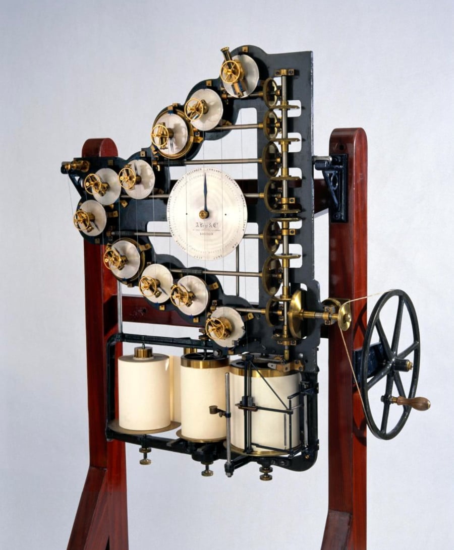 A photo of a mechanism that has 10 gears arranged in a V shape, with the V pointing to the left. In the center of the V is large round dial with a hand pointing to 12 o'clock. On the right hand side of the device there is a large hand crank. Several brass gears connect all the parts of the mechanism together. At the bottom, there appear to be three rolls of paper that record the results of the machine.