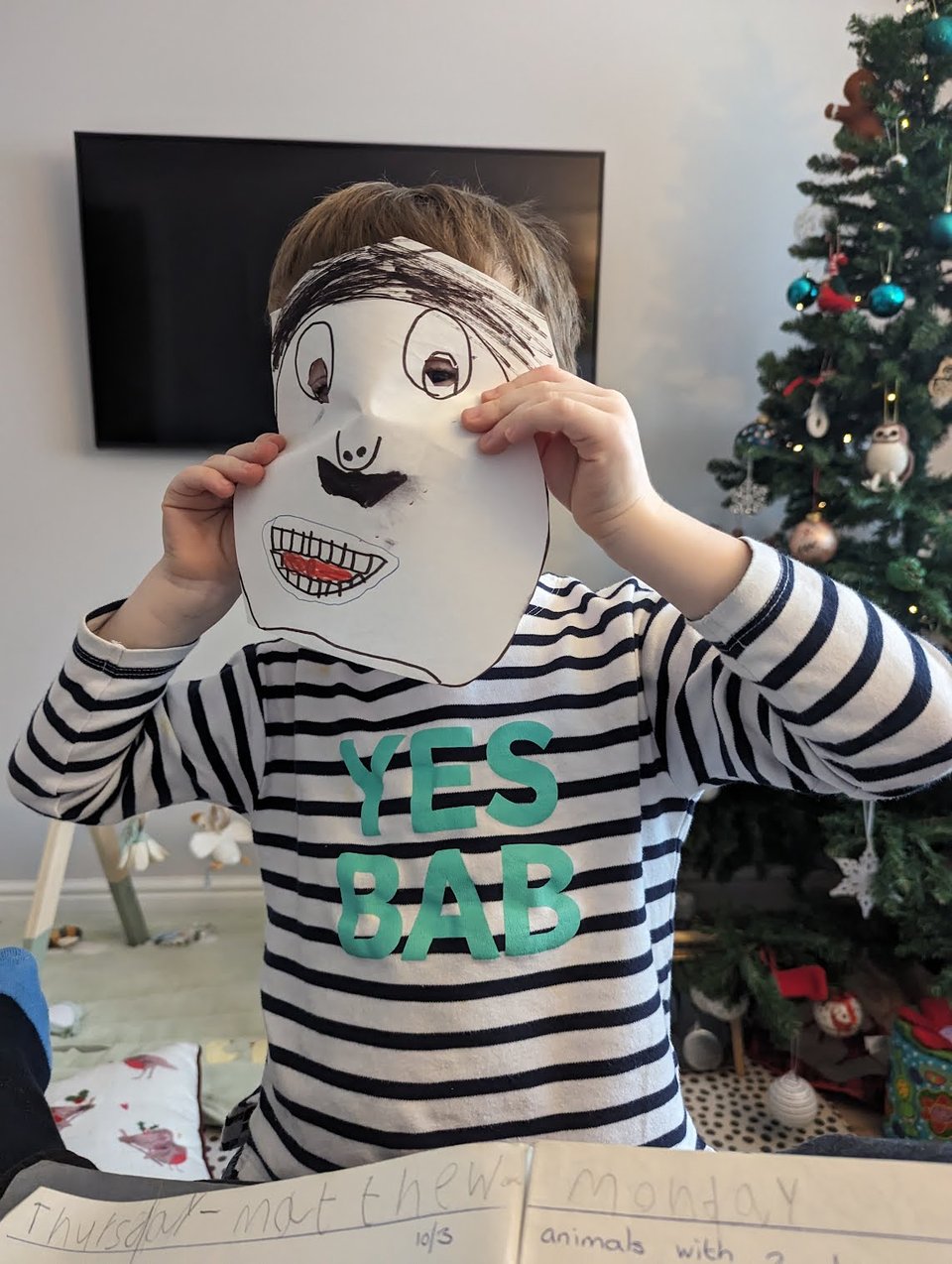 A child holding a paper mask of a man's face with prominent teeth and a moustache