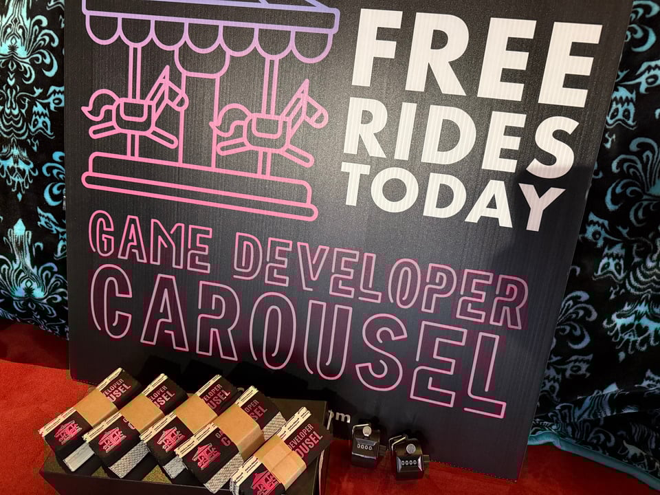 A sign with the Game Developer Carousel logo, word mark, and "Free Rides Today" rest on a wall. Bundles of Game Developer Carousel badge ribbons line the bottom, along with two handheld tally counters.