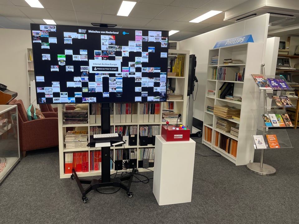 A large monitor is on a display in a museum lobby filled with books. There is a pedestal with a small red box on top. The monitor displays a grid of screenshots of webpages.